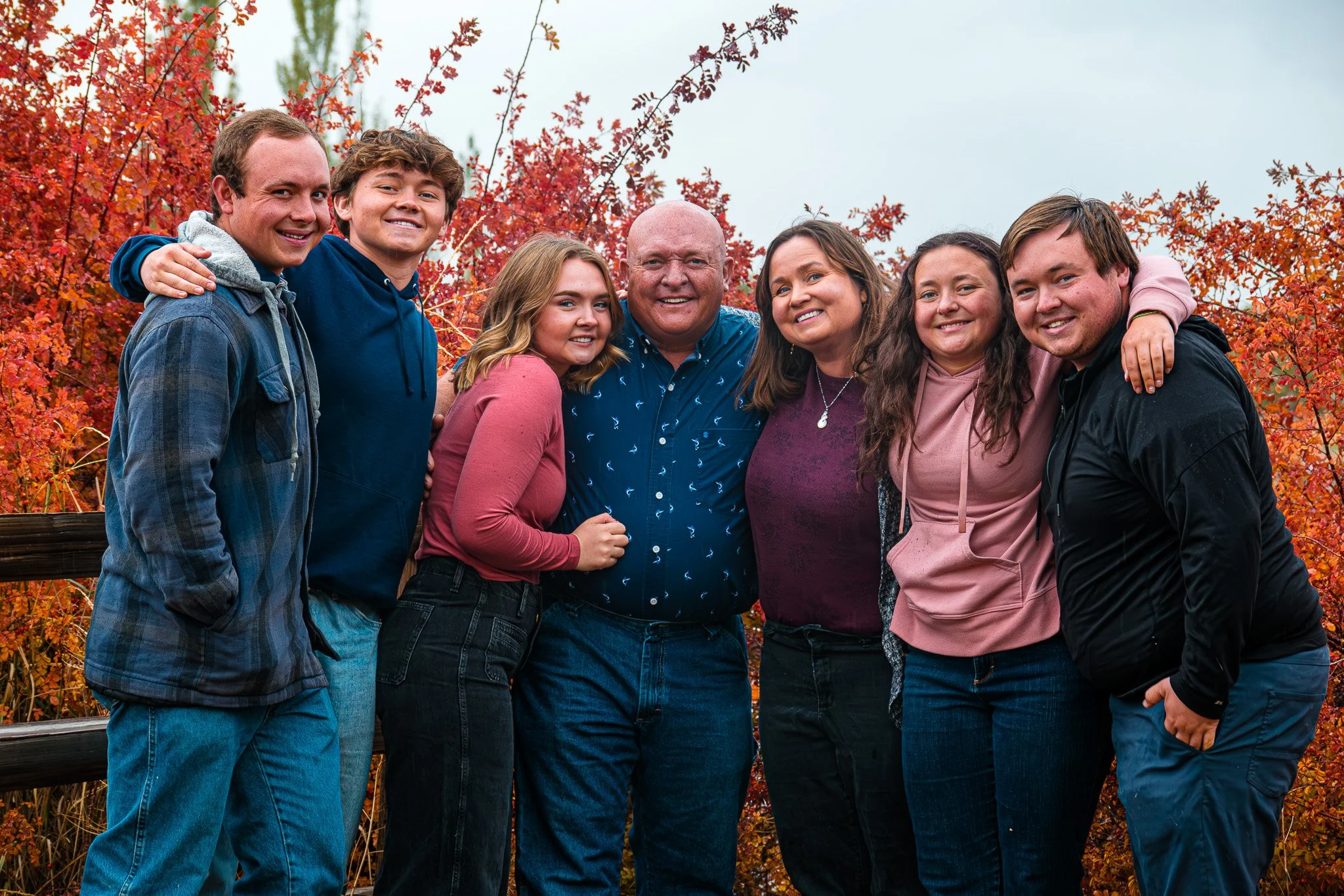 Family of seven in fall outdoor setting, standing close together, smiling, with colorful autumn foliage in the background.