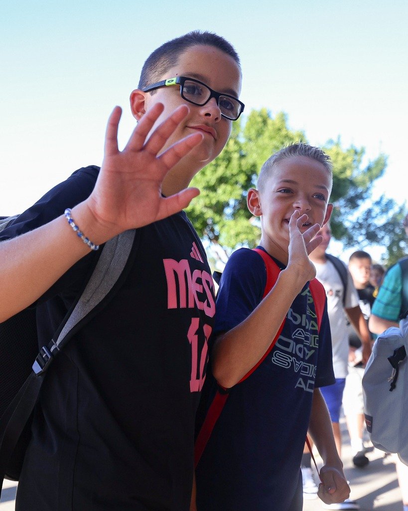 Two boys with backpacks waving and smiling outdoors, with a group of children and trees in the background.