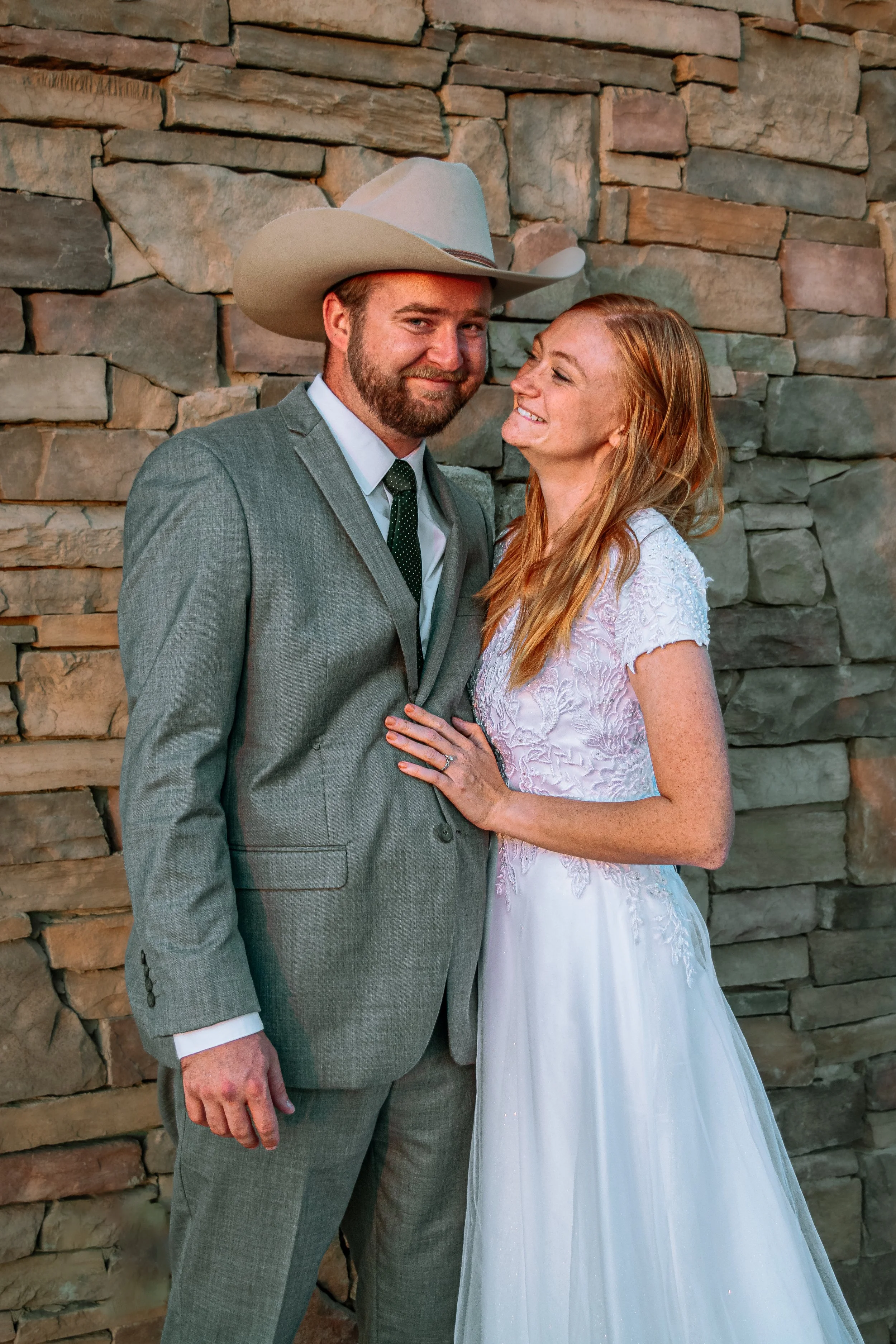 A newlywed couple stands close, smiling, with a stone wall backdrop. The groom is in a gray suit and cowboy hat, and the bride is in a white wedding dress with lace details, showing her ring and freckles.