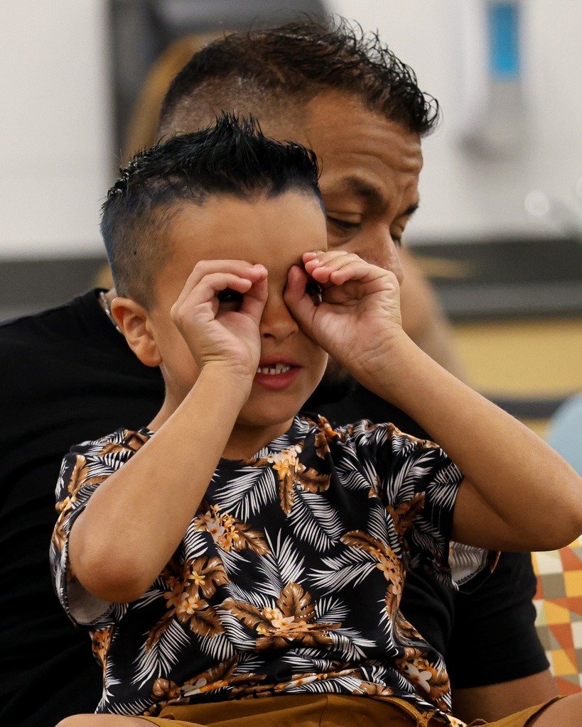 A young boy with a patterned shirt making a playful gesture with his hands around his eyes, sitting on an adult's lap in an indoor setting.