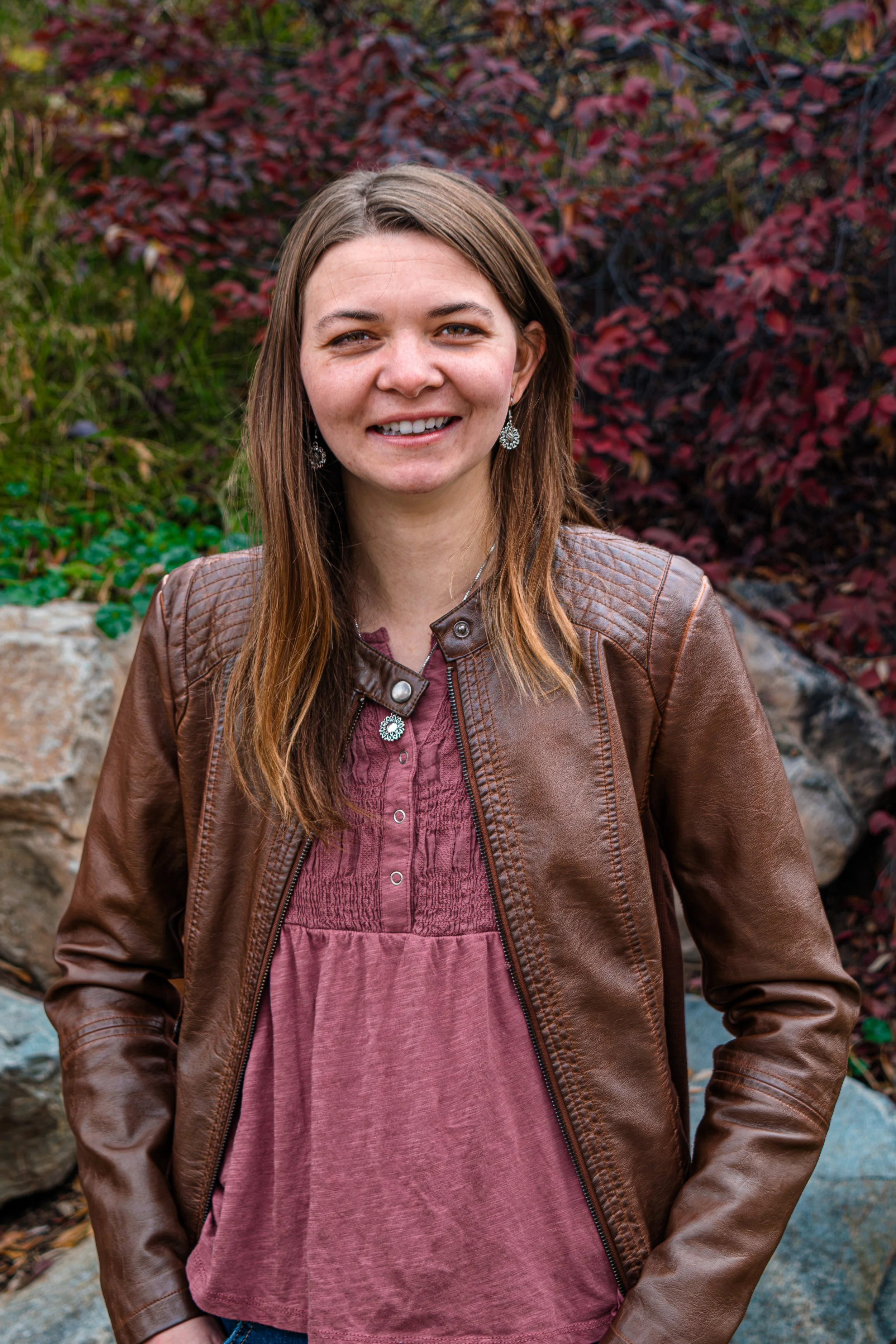 A woman with long brown hair and light skin smiling outdoors, wearing a brown leather jacket, earrings, and a red blouse, with red and green foliage in the background.