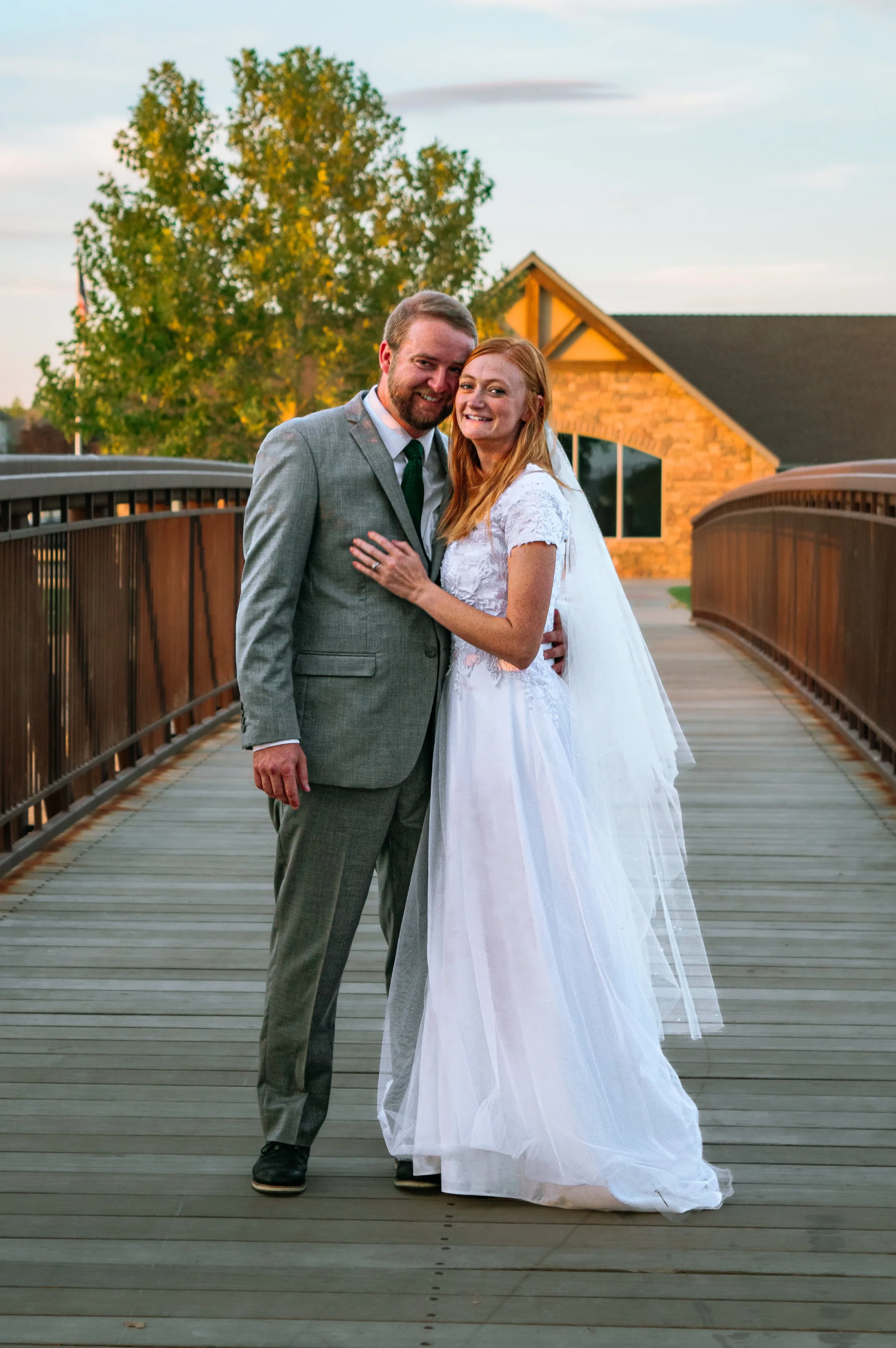 A bride and groom in wedding attire standing on a wooden bridge during sunset, smiling at the camera.