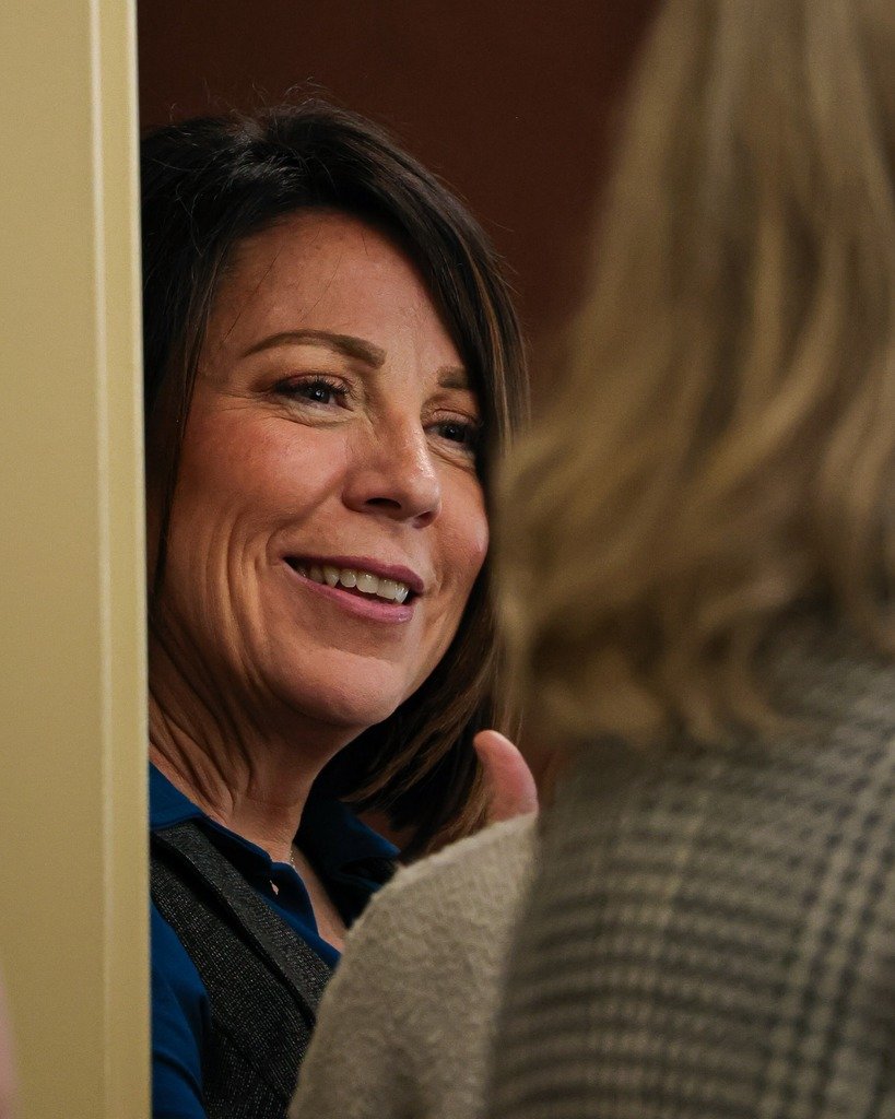 A woman with short dark hair smiling and talking to another person with blonde hair, seen from behind, in an indoor setting.