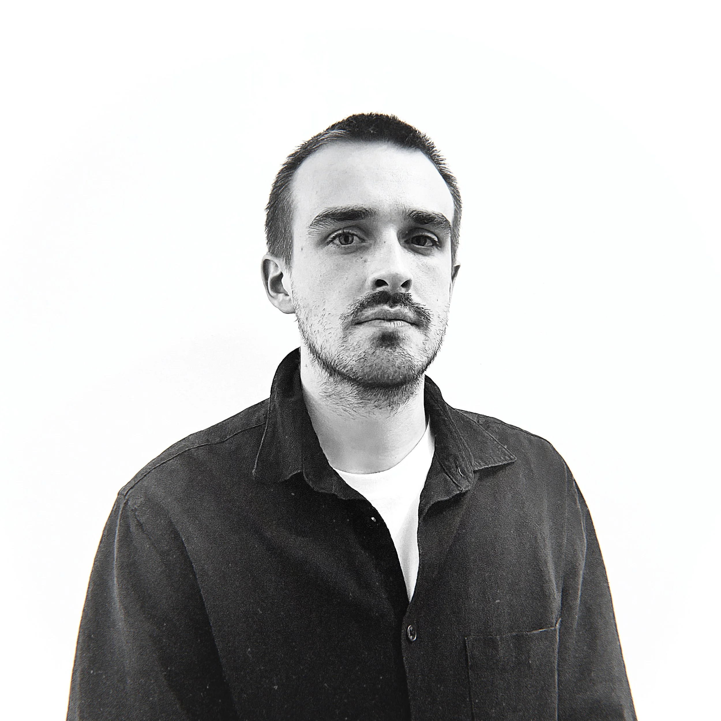 Black and white portrait of a young man with short hair, facial hair, wearing a dark button-up shirt and a light-colored undershirt, against a plain white background.