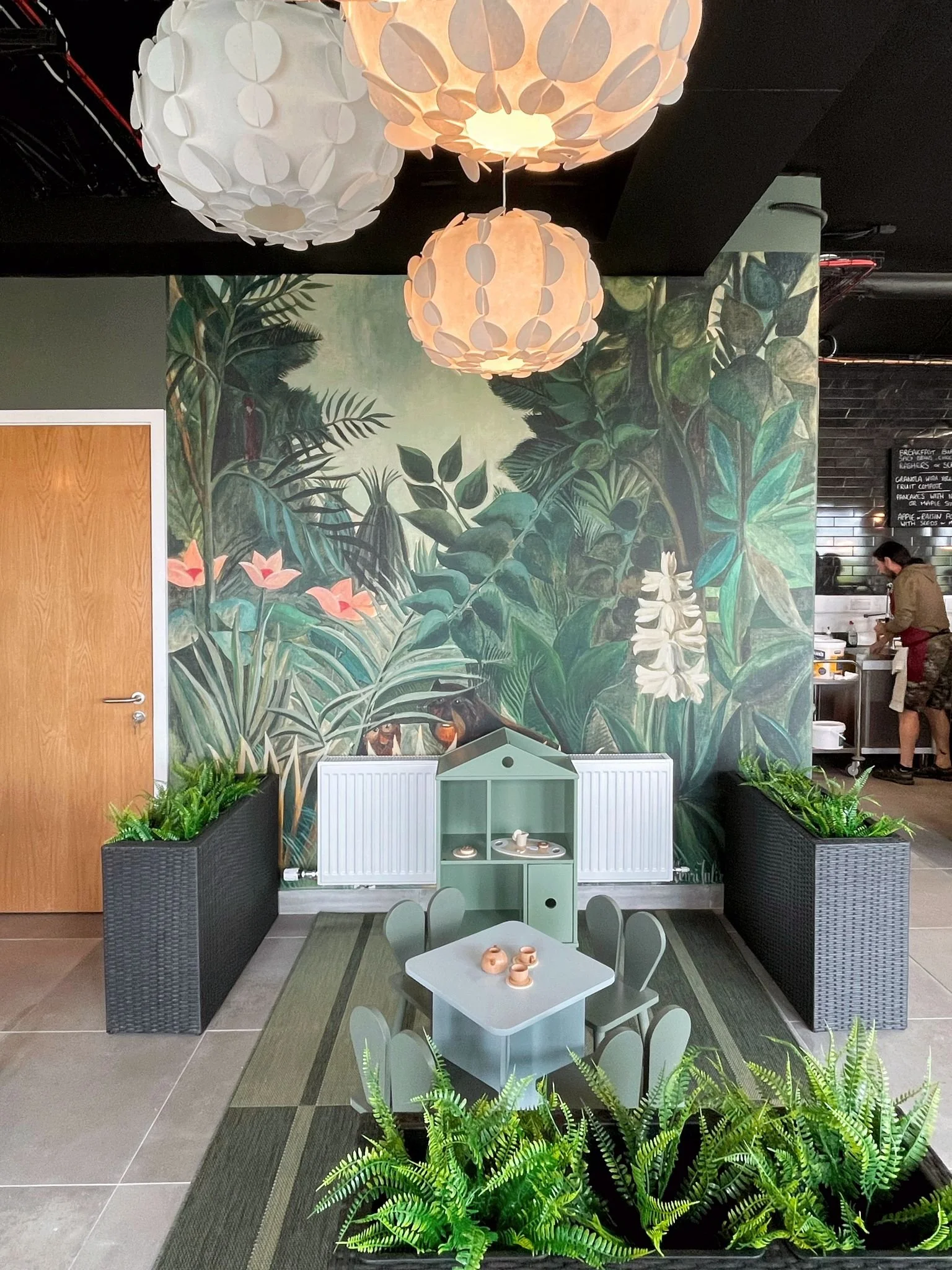 Cafe seating area with jungle-inspired wallpaper, striped flooring, pendant lights, and a playful family-friendly layout.