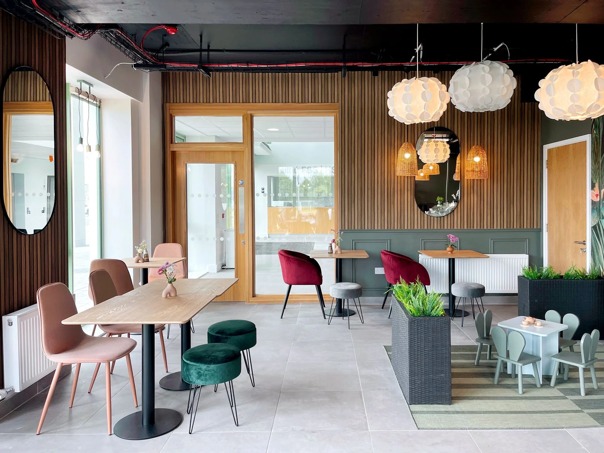 Light-filled dining area with a communal table, velvet seating, warm wood finishes, and garden views.
