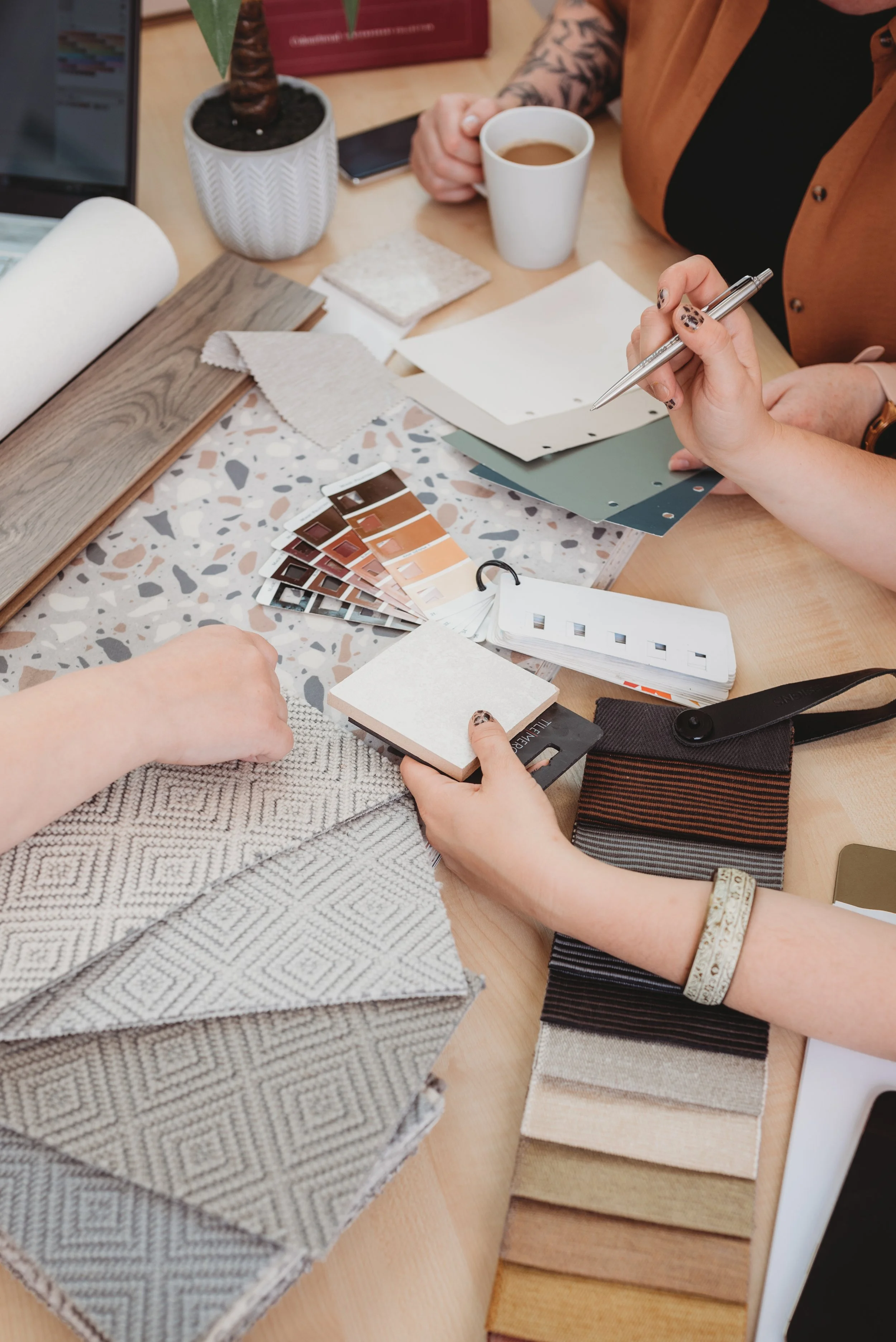People discussing interior design samples, fabric swatches, and paint color samples on a table, with a laptop, potted plant, and a cup of coffee also visible.