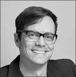 A black and white portrait of a man with glasses, short dark hair, and a broad smile, wearing a dark blazer.