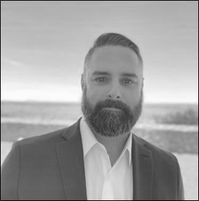 A man with a beard and short hair, wearing a suit jacket and white shirt, standing outdoors with a blurred landscape in the background.