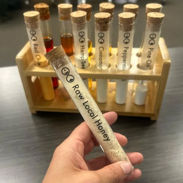 Test tube filled with a powder labeled 'Raw Local Honey' held by a person's hand, with a wooden test tube rack containing similar test tubes of various spices and powders in the background.