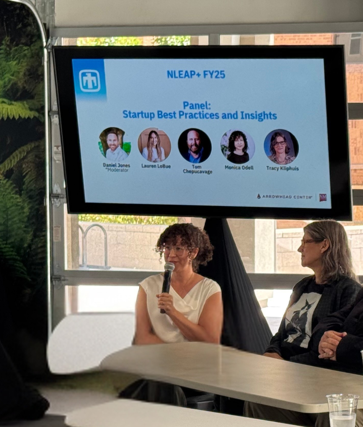 A woman sitting at a table with a microphone, speaking during a panel discussion. Behind her, a large screen displays the panel title 'Startup Best Practices and Insights' with photos and names of five panelists, including Daniel Jones as moderator, Lauren LoBue, Tom Chepucavage, Monica Odell, and Tracy Kliphuis. The setting appears to be an indoor event space with large windows.