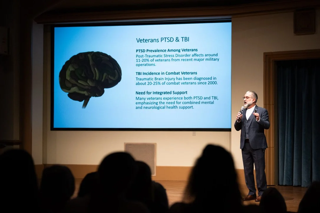 A man in a dark suit and glasses giving a presentation on PTSD and TBI among veterans. The slide shows a graphic of a brain and text about prevalence, incidence, and support needs.