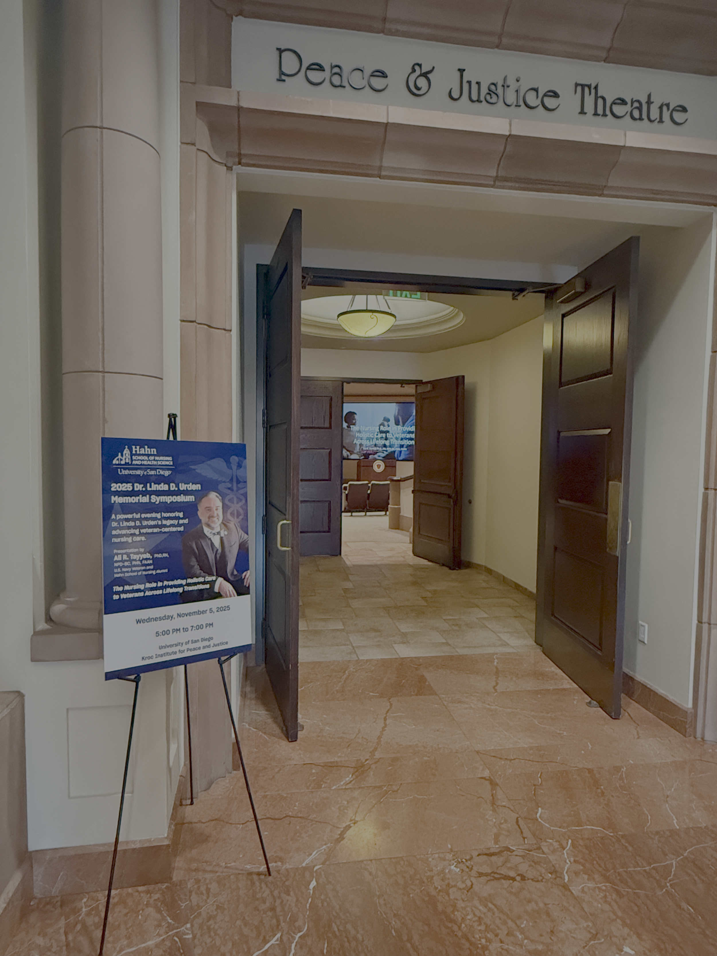 Entrance to Peace & Justice Theatre with open doors, wooden panels, and a sign for a symposium on nursing and veteran care.