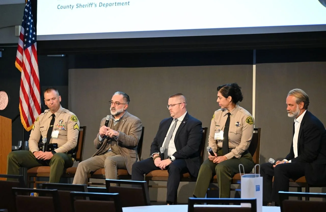 Panel discussion with five people seated on stage, including law enforcement officers in uniform and civilians, in front of a large screen displaying 'County Sheriff's Department' and an American flag on the left.