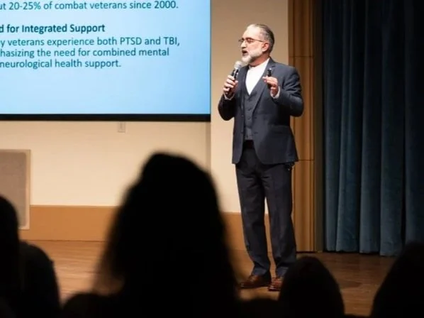 Speaker presenting on stage at a conference with a podium and presentation slide visible, audience in foreground.