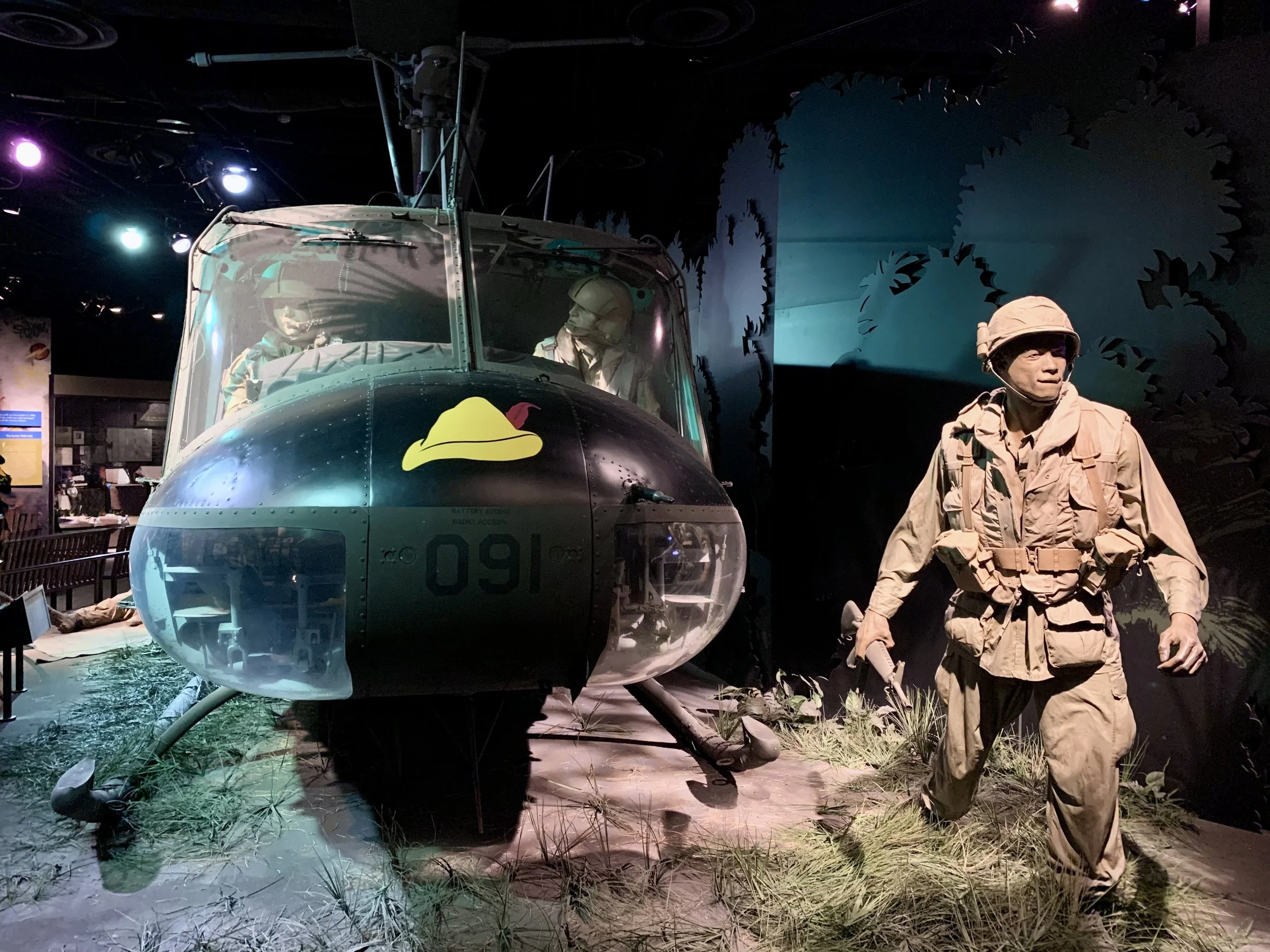 Smithsonian Museum depicting a Huey Helicopter on the ground with two pilots in a mud and grassy area of vietnam during the Vietnam war with another military service member in green BDU's standing to the side.