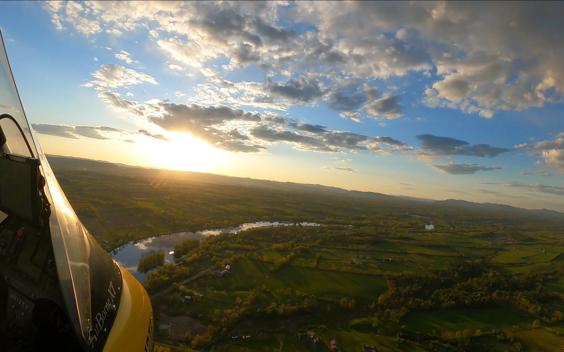 A scenic view of a river winding through green fields and trees during sunset, taken from inside a helicopter cockpit visible on the left side of the image.