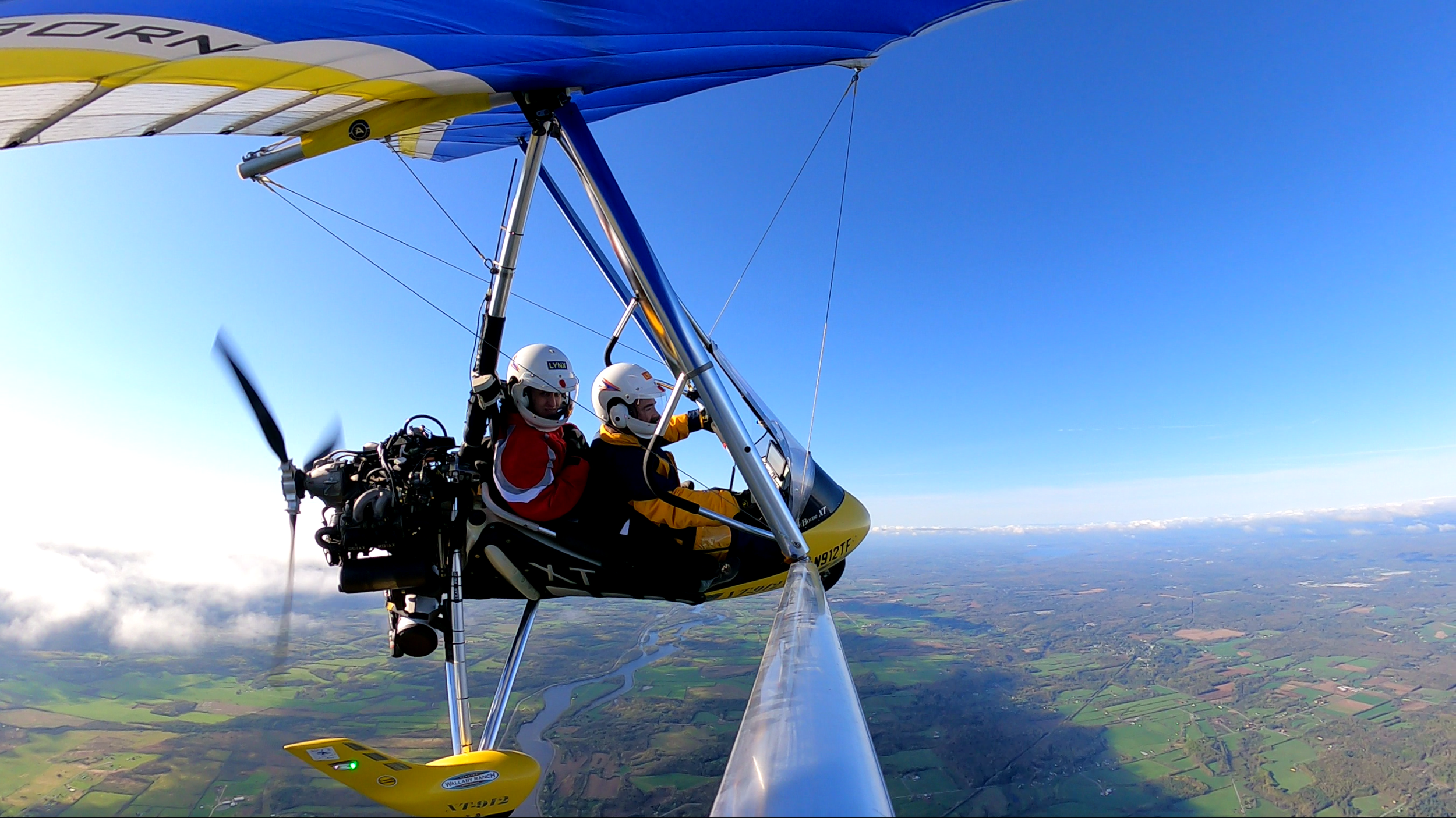 An ultralight aircraft flying high above the countryside with two people inside, one wearing a red jacket and the other a yellow jacket, both with helmets.