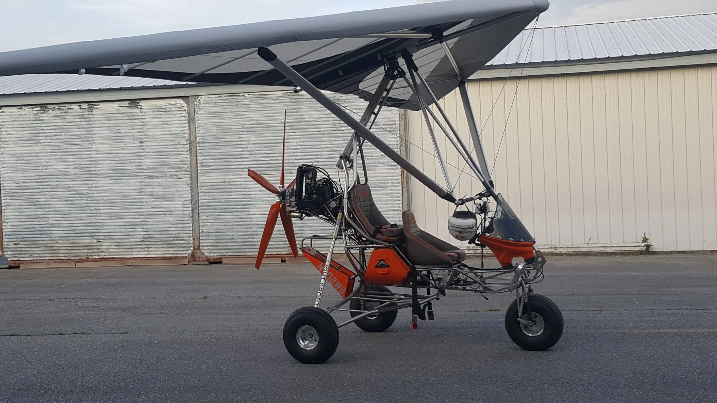 A small autogyro aircraft with a single seat, orange and black accents, and a large rotor on top, parked on an asphalt surface near a weathered metal building.