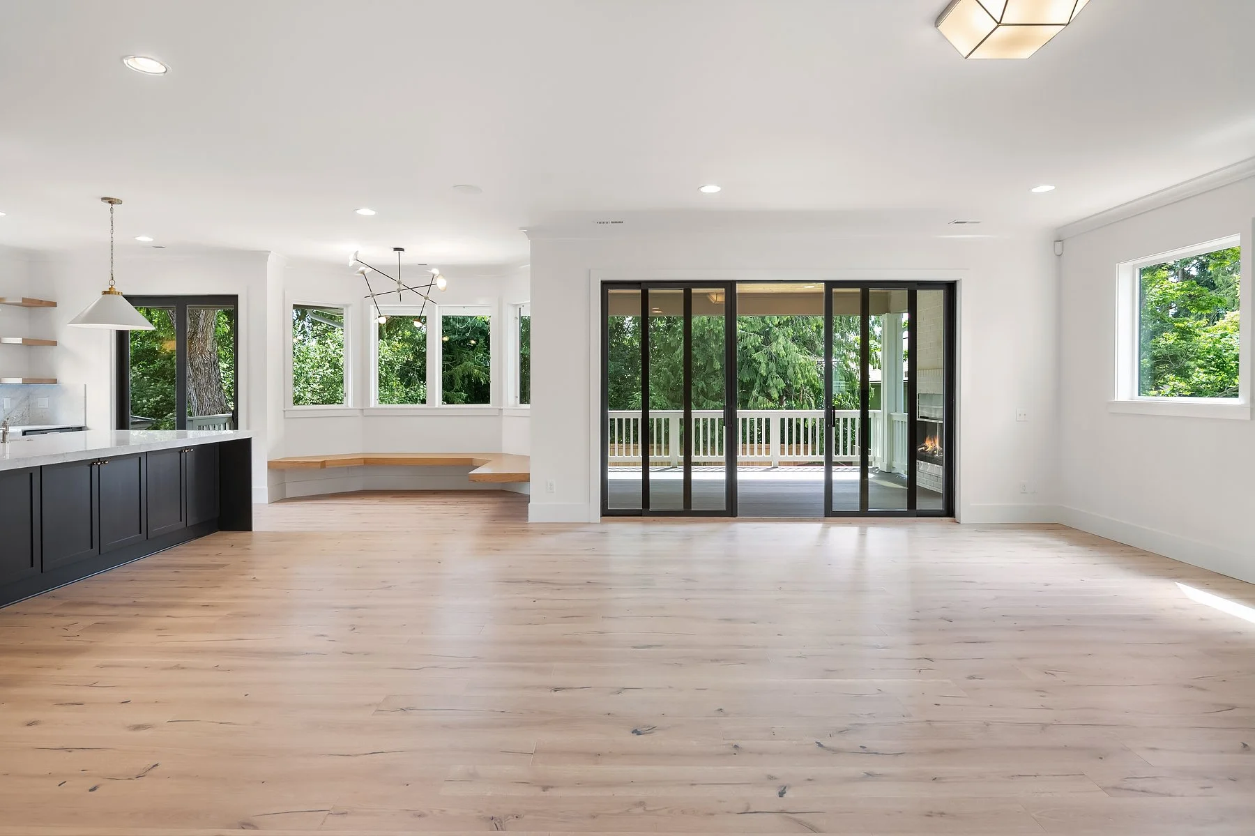 Empty living room with light wood flooring, white walls, large windows, and sliding glass doors leading to a balcony with trees outside.