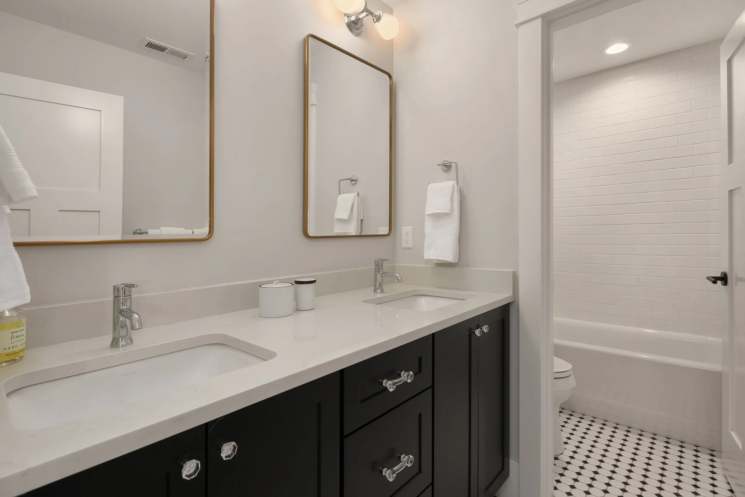 Bathroom with double vanity, two mirrors, black cabinets, and a shower area with white brick wall tiles