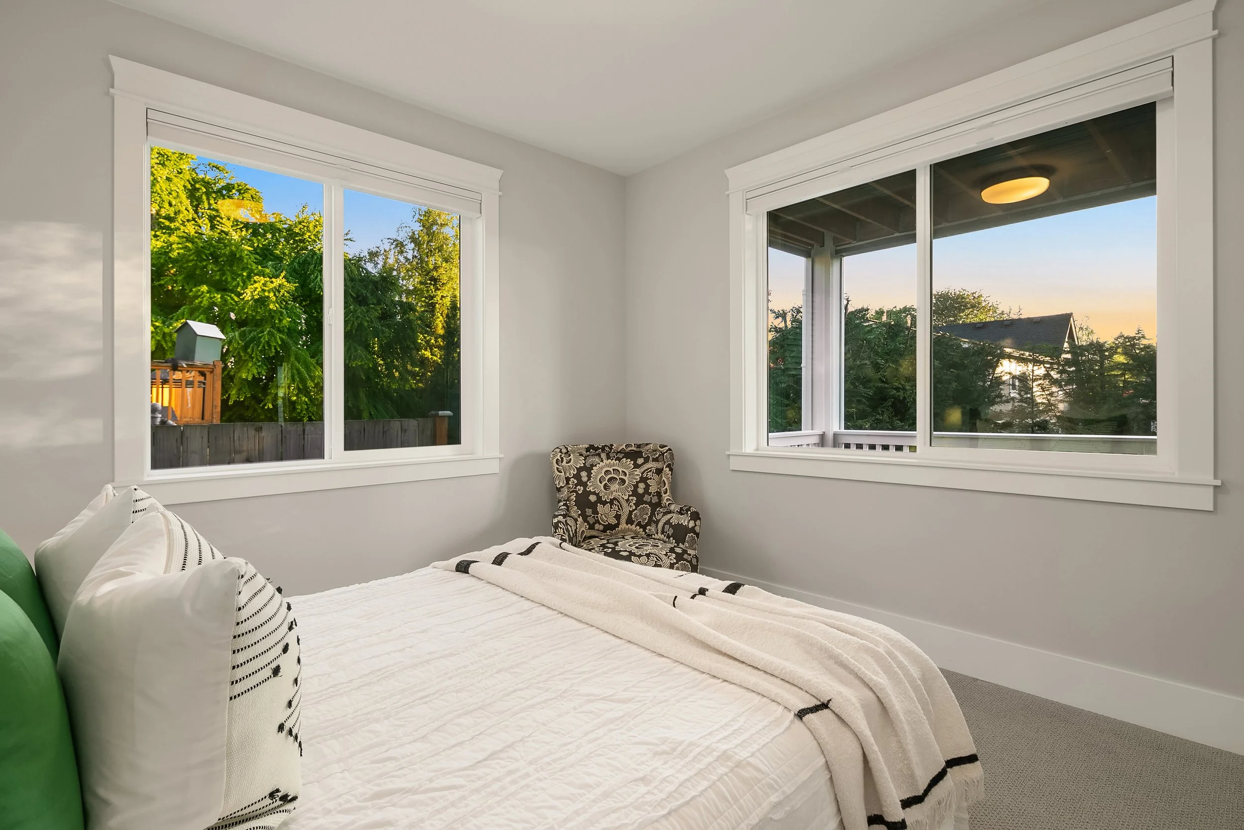 A bedroom with white walls, two large windows showing a green yard outside, a bed with white bedding and pillows, and a patterned armchair in the corner.