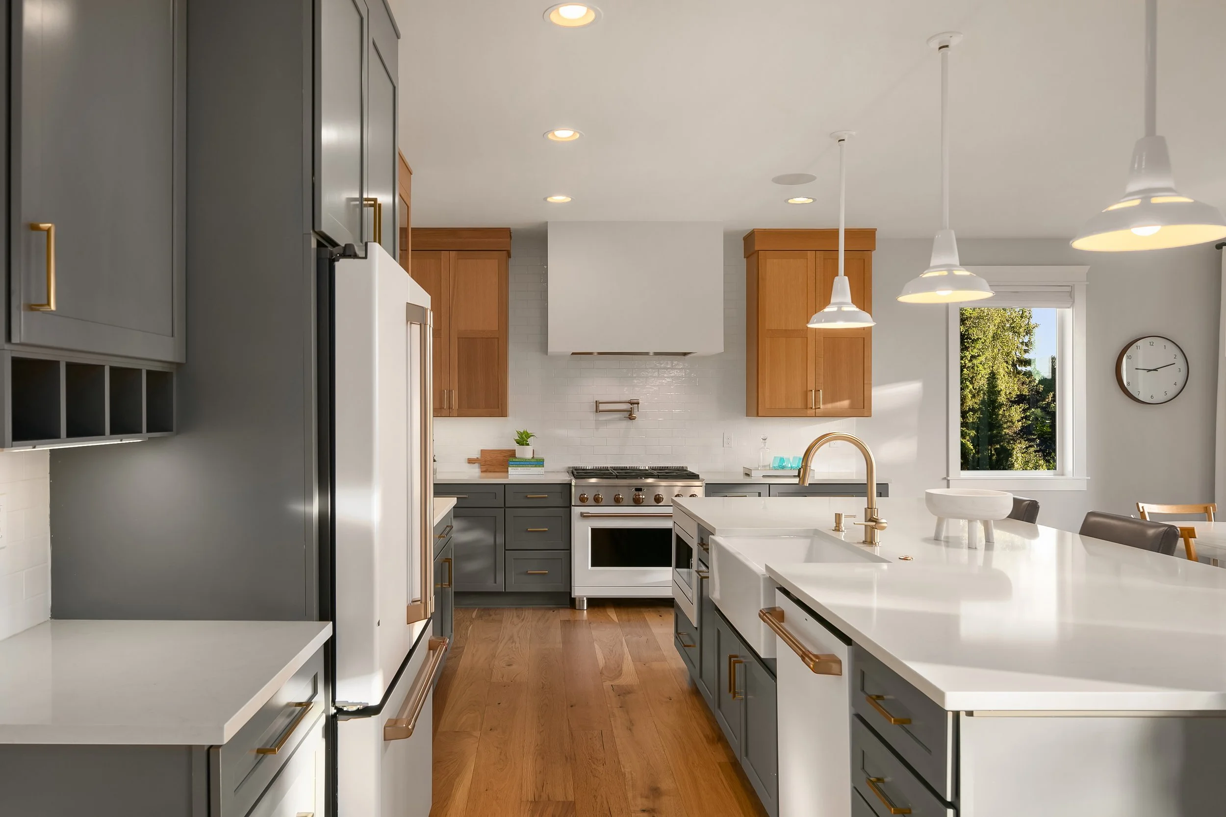 Modern kitchen with gray and wooden cabinets, white countertop, stainless steel appliances, and a large window showing greenery outside.