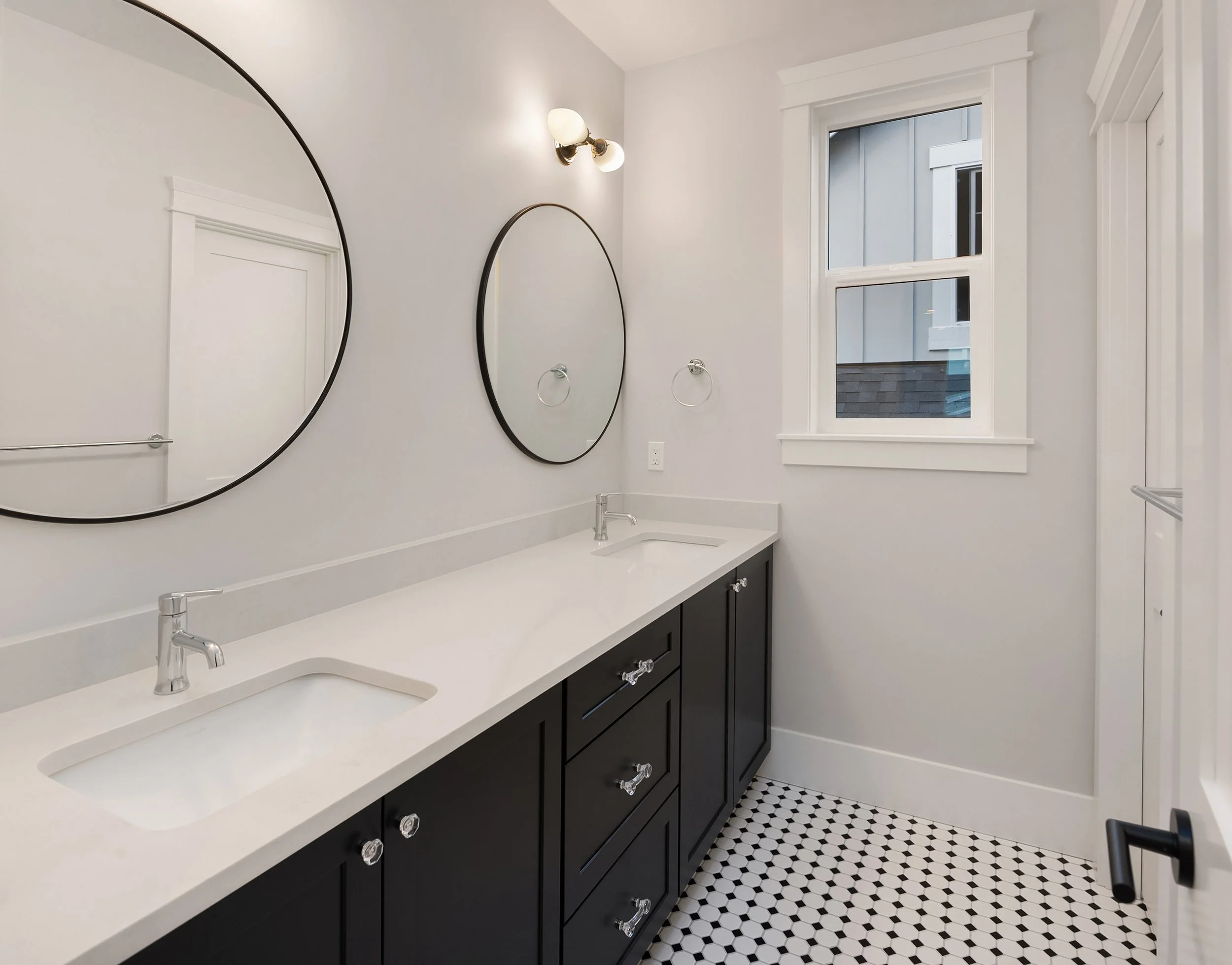A modern bathroom with two oval mirrors above a white countertop with two sinks, black cabinets, a window, and black and white tiled floor.