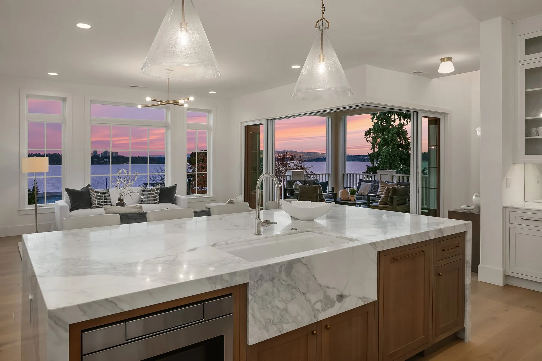 Modern kitchen with white marble island, large windows with a view of a lake and sunset, and a living area with a sofa and balcony.