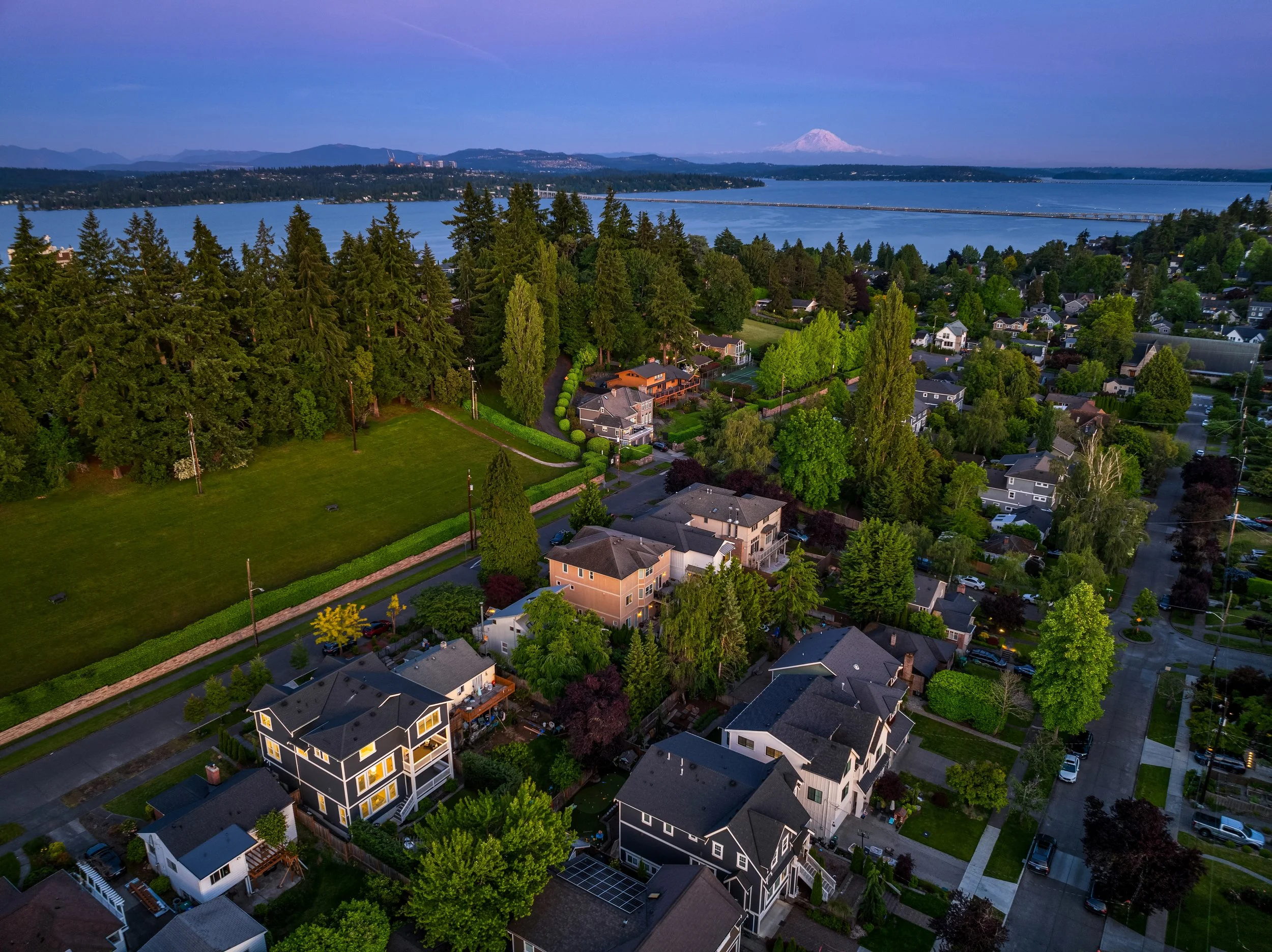 Aerial view of a suburban neighborhood with houses, trees, a park, a lake, and mountains in the background during dusk or dawn.