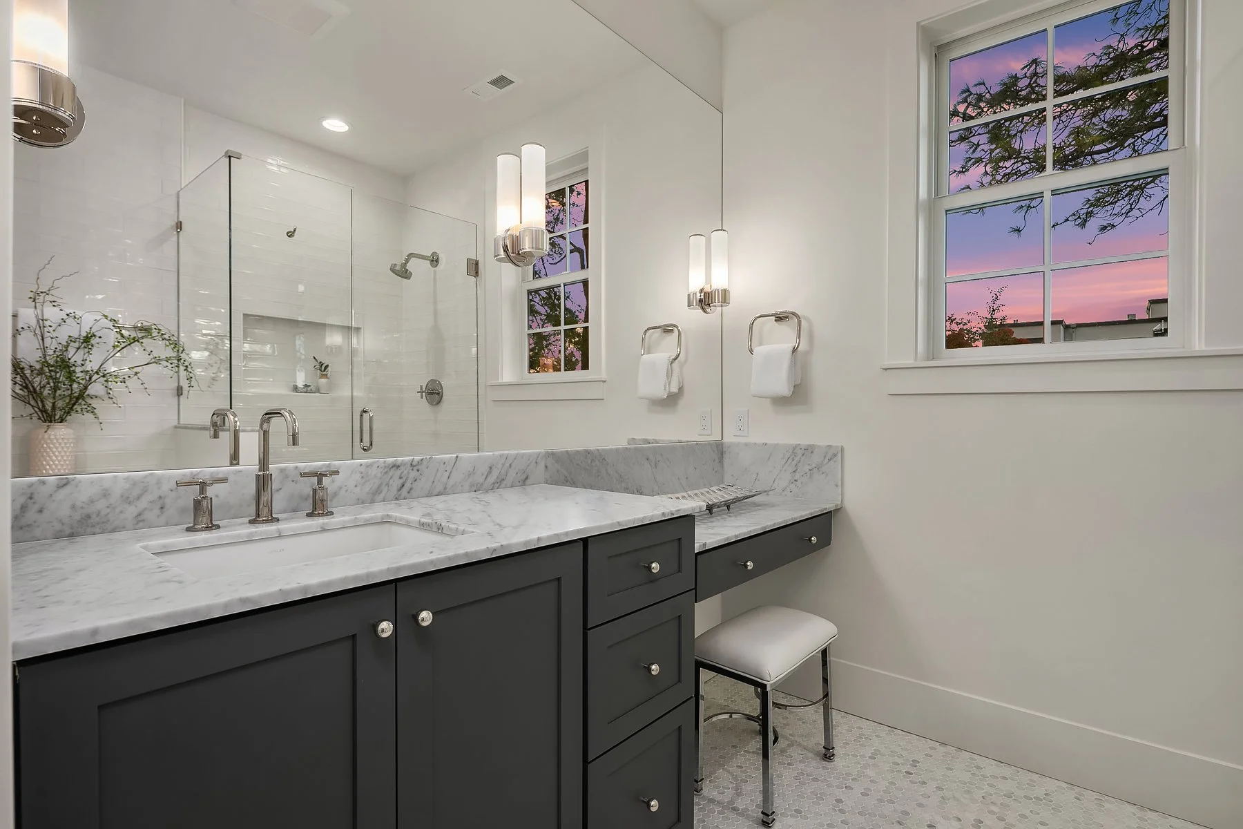 Modern bathroom with marble countertop, navy cabinets, large mirror, and window showing a sunset sky.