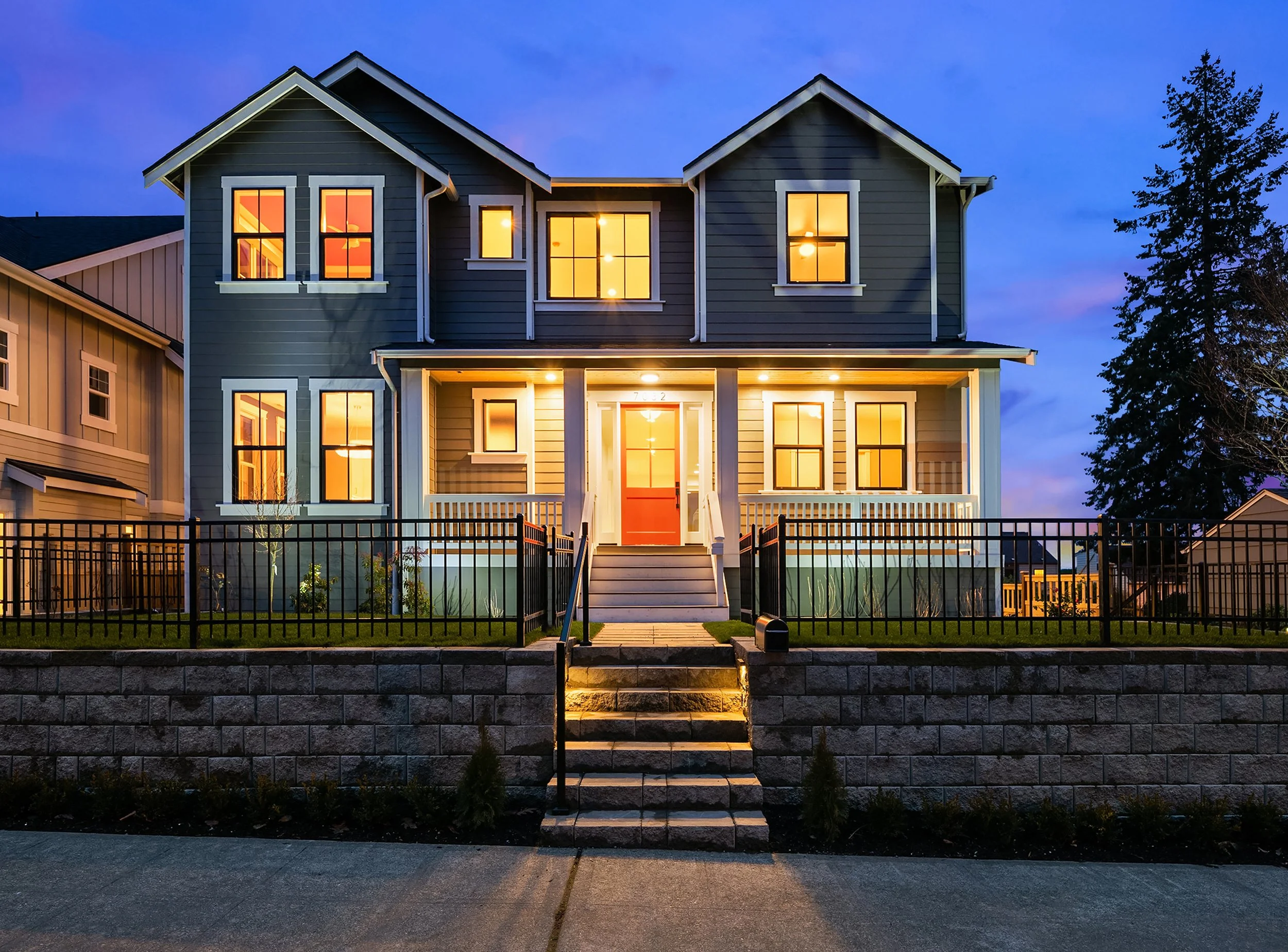 A two-story house with a dark gray exterior, illuminated windows, and a red front door, with a front yard enclosed by a black metal fence and stone steps leading up to a porch during evening hours.