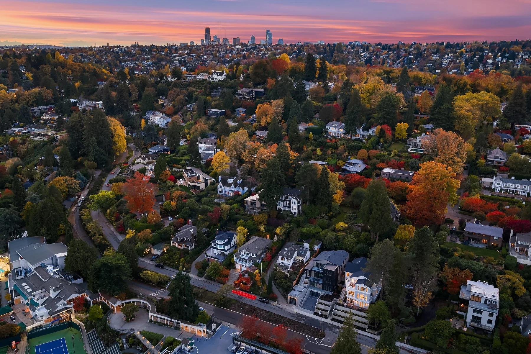 Aerial view of a neighborhood with colorful autumn trees and houses, with a city skyline in the distance under a sunset sky.