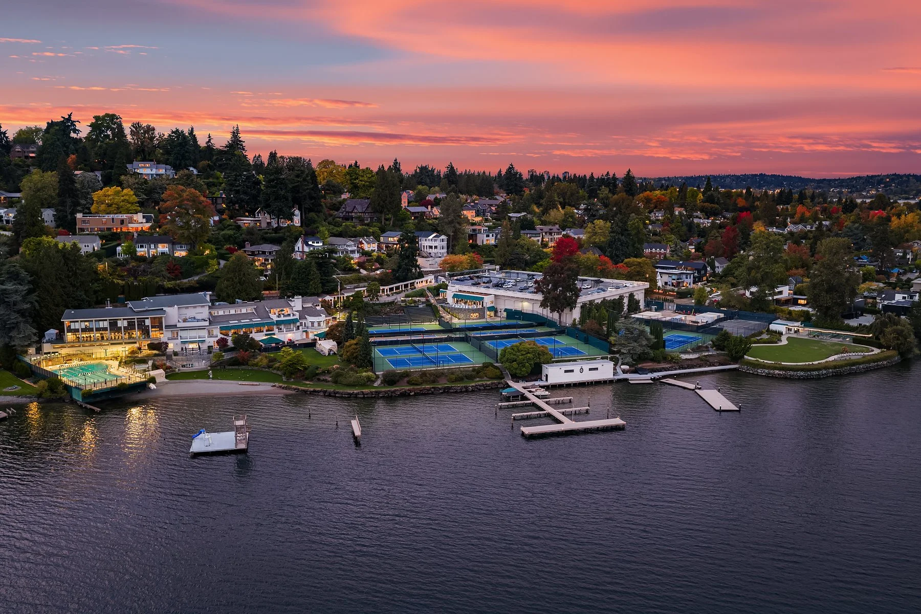 An aerial view of a residential neighborhood by a lake at sunset, featuring tennis courts, a clubhouse, and various houses with trees showing autumn colors.
