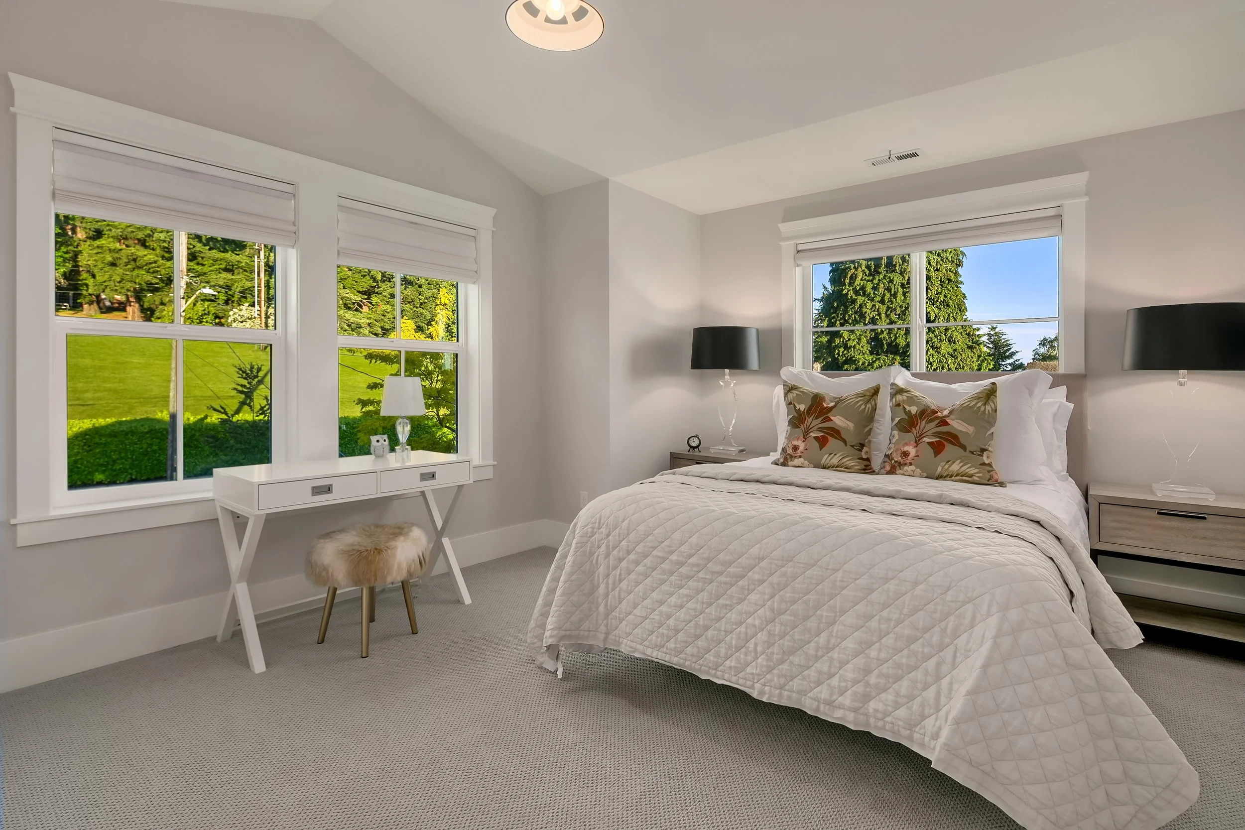 A bright bedroom with white walls, three large windows showing green trees outside, a bed with white quilt and two floral pillows, two black lamps on nightstands, a small desk with a lamp and small owl figurines, and a fluffy stool nearby.