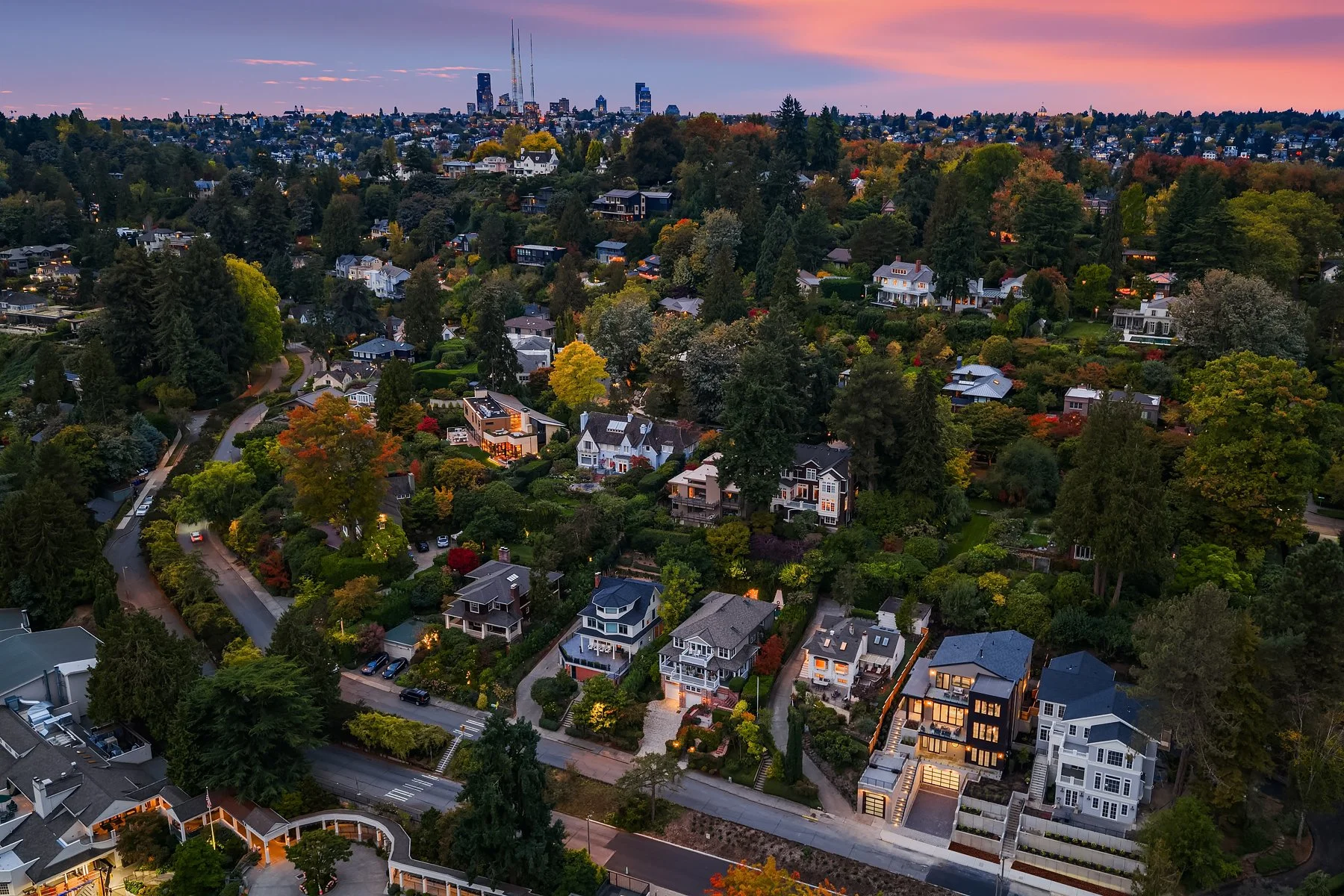Aerial view of a residential neighborhood surrounded by trees at dusk, with a city skyline in the background and colorful sunset sky.