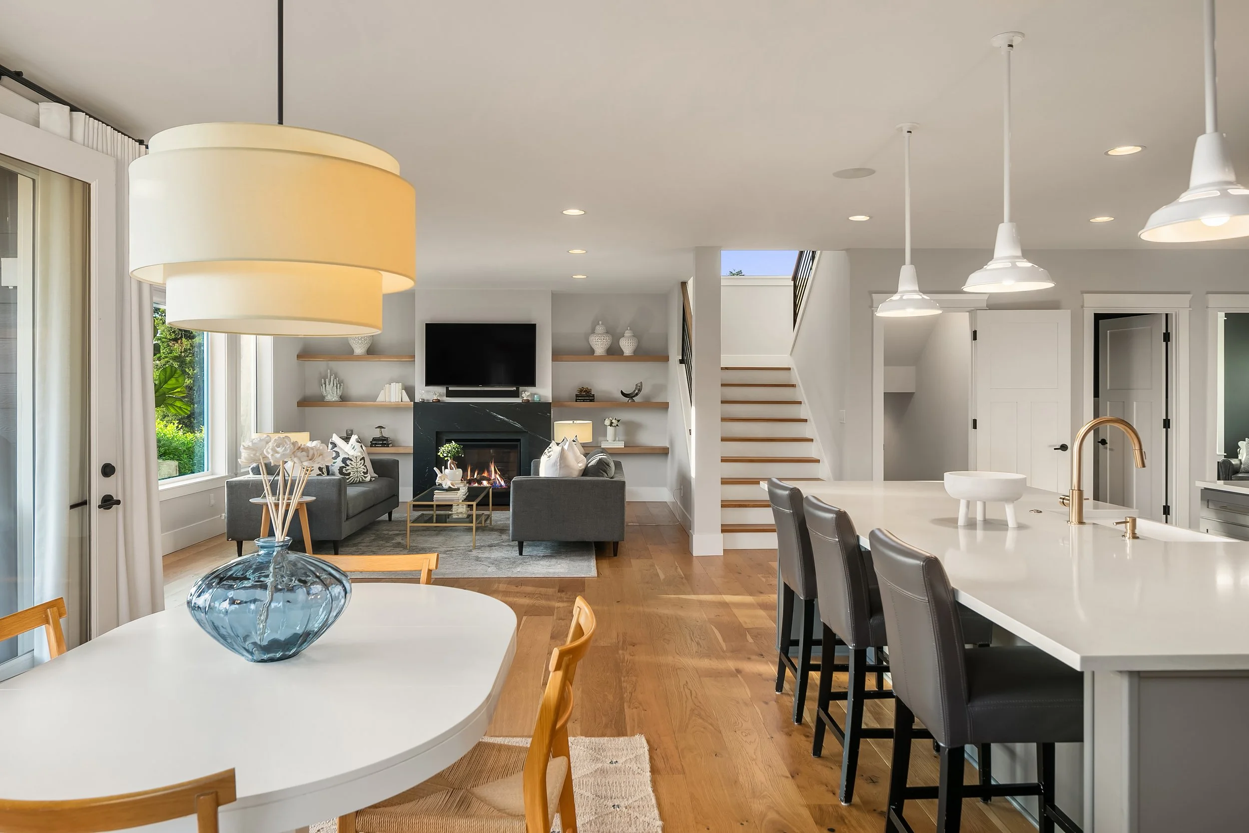 A modern open-concept living room and kitchen with white walls and wooden floors, gray sofas in the living room with a fireplace, a wall-mounted TV, and built-in shelves. The kitchen has a large white island with a brushed gold faucet, black barstool