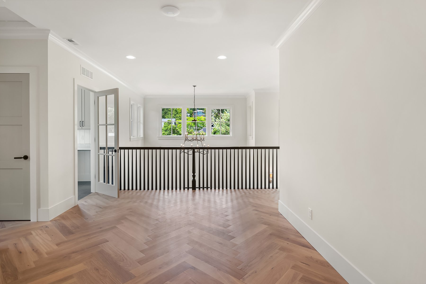 Empty living room with white walls, light wood herringbone floor, black railing, and large windows with greenery outside.