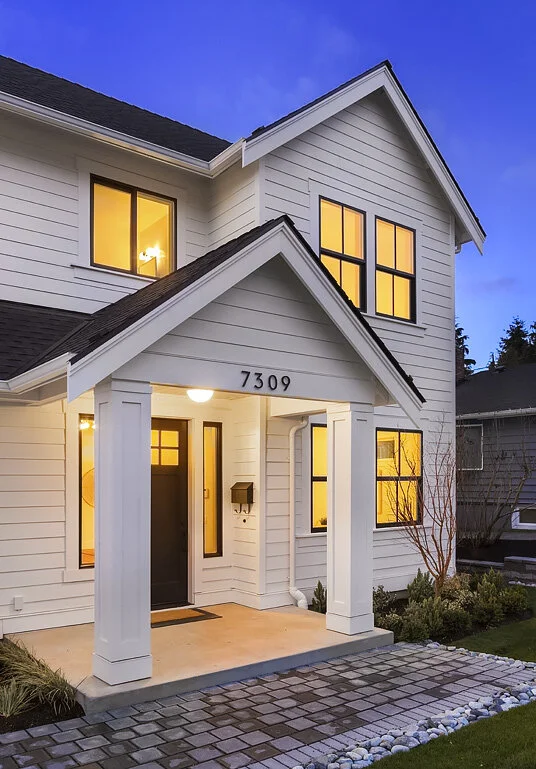 Two-story white house with black front door, illuminated windows, stone pathway, and landscaped yard during dusk.