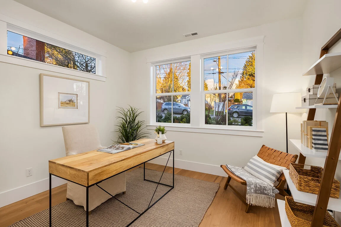Bright home office with a wooden desk, beige chair, and cozy lounge chair with a striped pillow, white lamp, and shelves with books and baskets.