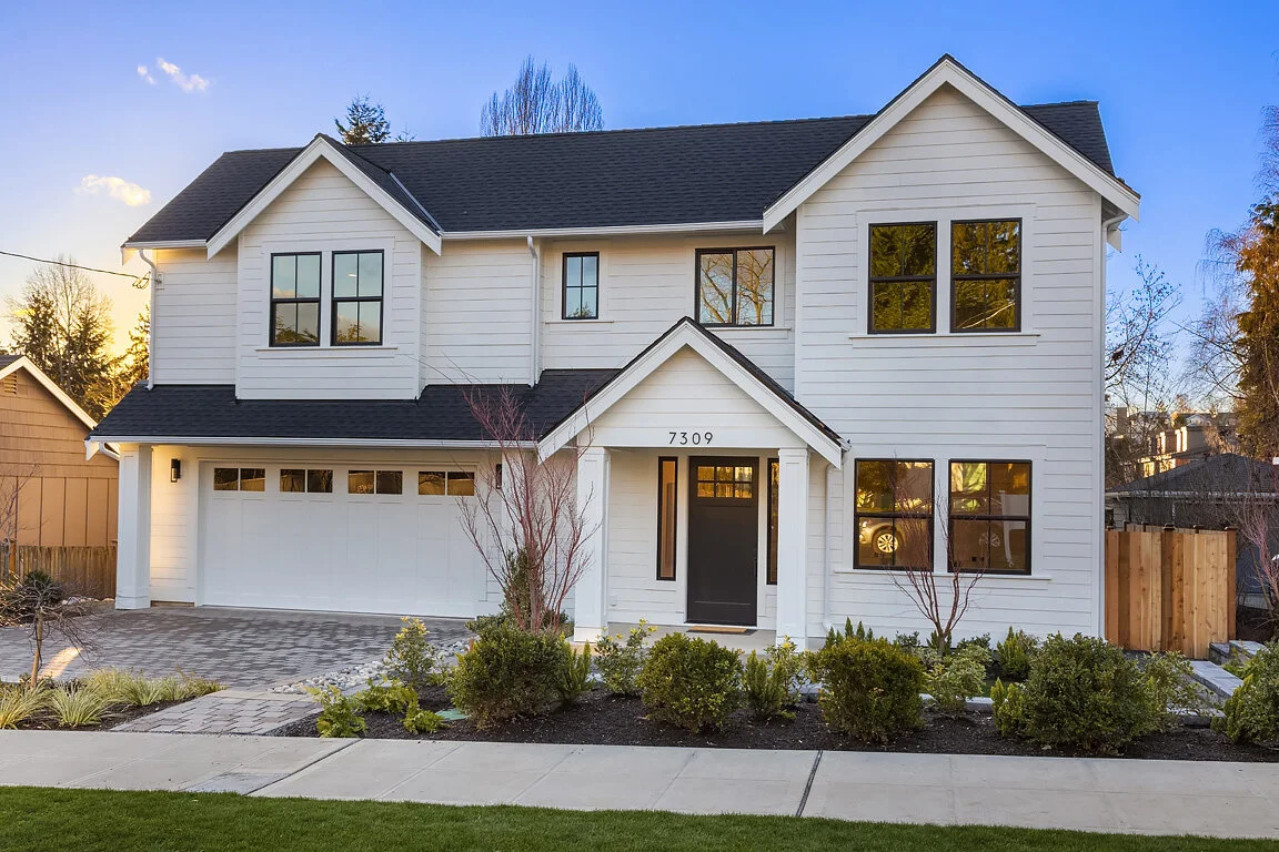 Modern two-story white house with black roof, large front windows, and a front porch with a black door, surrounded by a landscaped yard with shrubs and young trees.