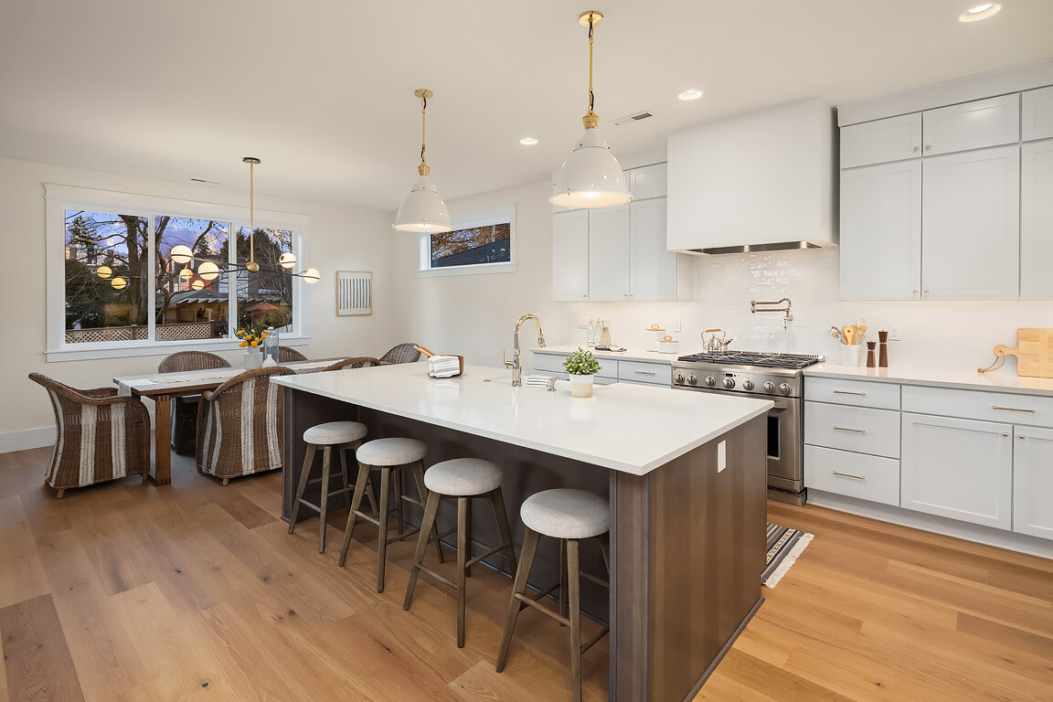 Modern kitchen with a large central island, white cabinetry, stainless steel stove, wooden floors, and a dining area with chairs near a window.