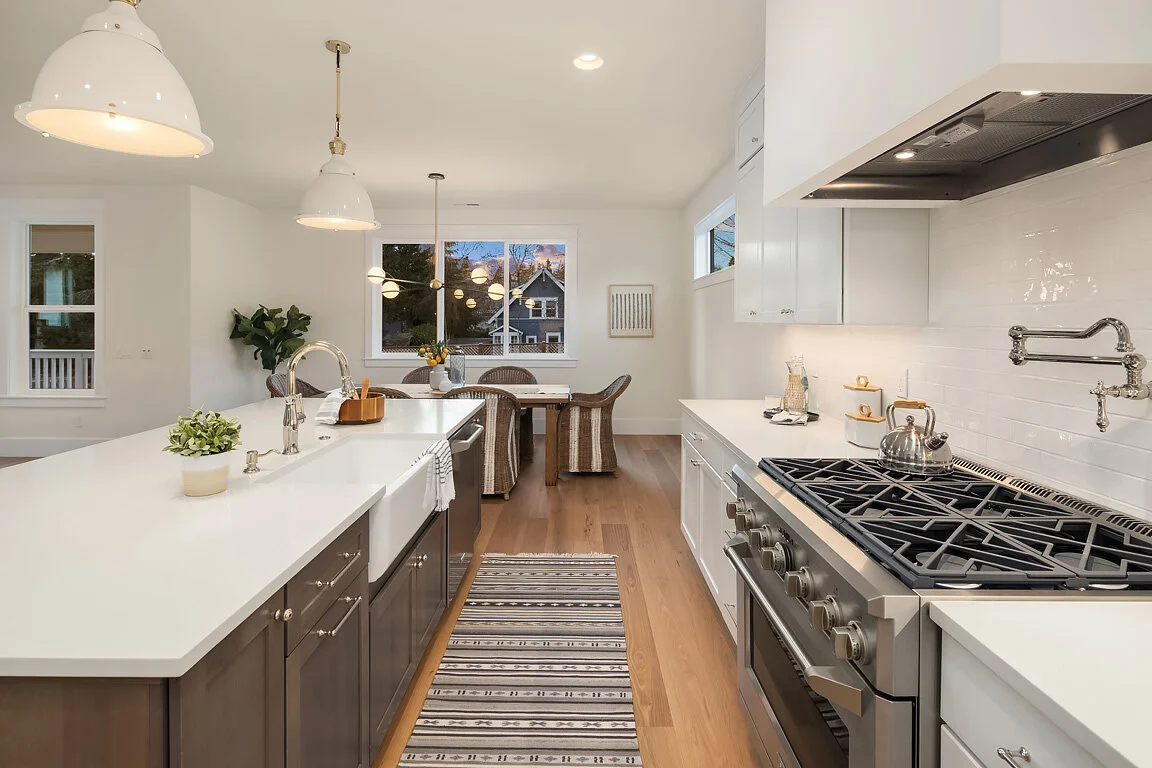 Modern kitchen with white and dark cabinetry, stainless steel oven, and light wooden floors. Kitchen island with sink and plants, dining table with chairs nearby, and large windows showing a house outside.