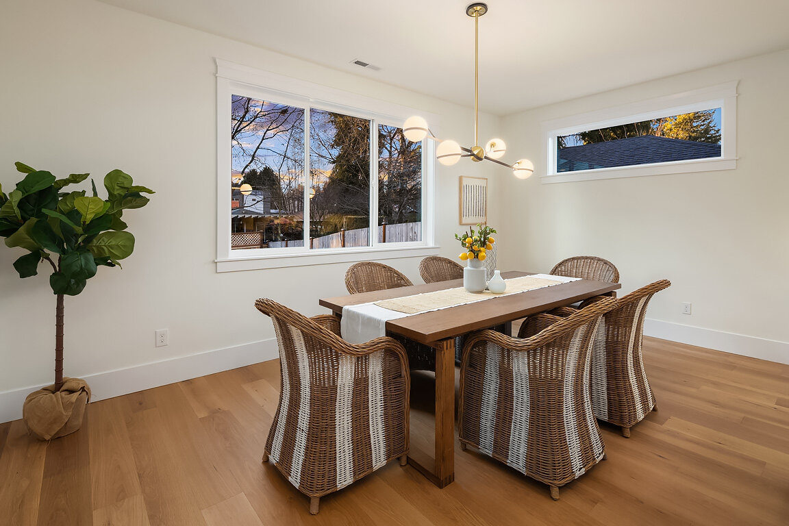 A dining room with a wooden table, six woven rattan chairs, a white table runner, a potted yellow flower, a small white vase, and a minimalist chandelier. Large window with a view of trees and a house in the background.
