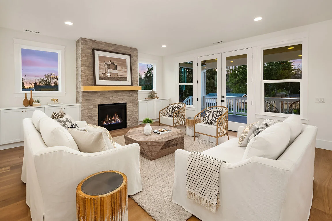 Living room with white sofas, patterned armchairs, wood coffee table, fireplace, and large windows with a view of a deck and trees at sunset.