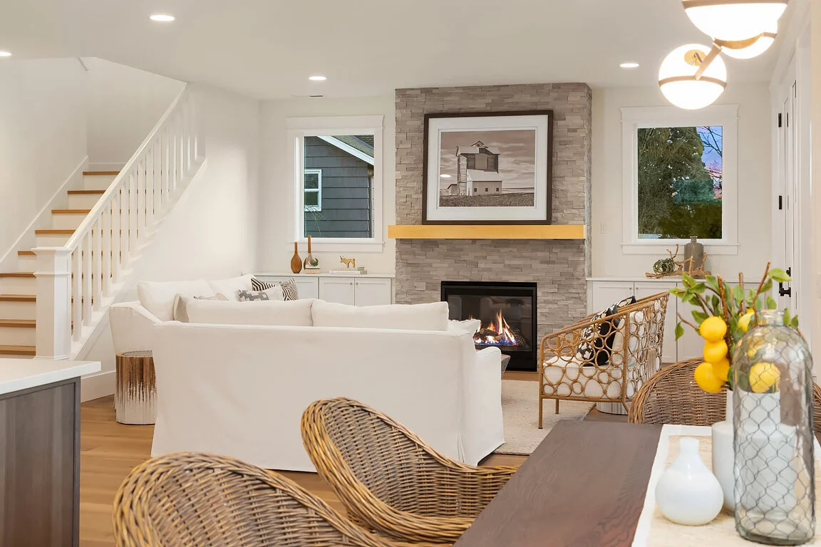 Living room with a stone fireplace, white sofas, wicker chairs, a wooden coffee table, and decorative vases, illuminated by recessed ceiling lights and a modern chandelier.