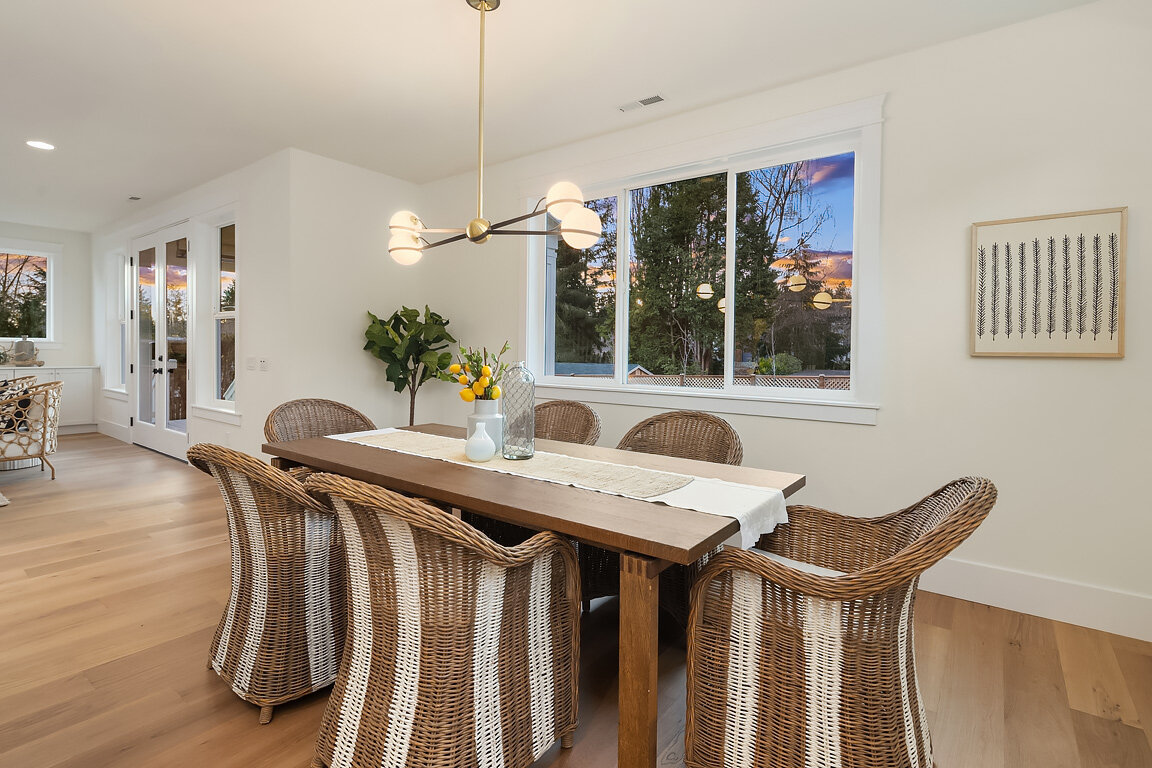 Image of a modern dining room with a wooden table, six woven wicker chairs, a white table runner, and a centerpiece with yellow lemons in a white vase. Large windows show trees and a sunset outside, and a contemporary chandelier hangs above the table