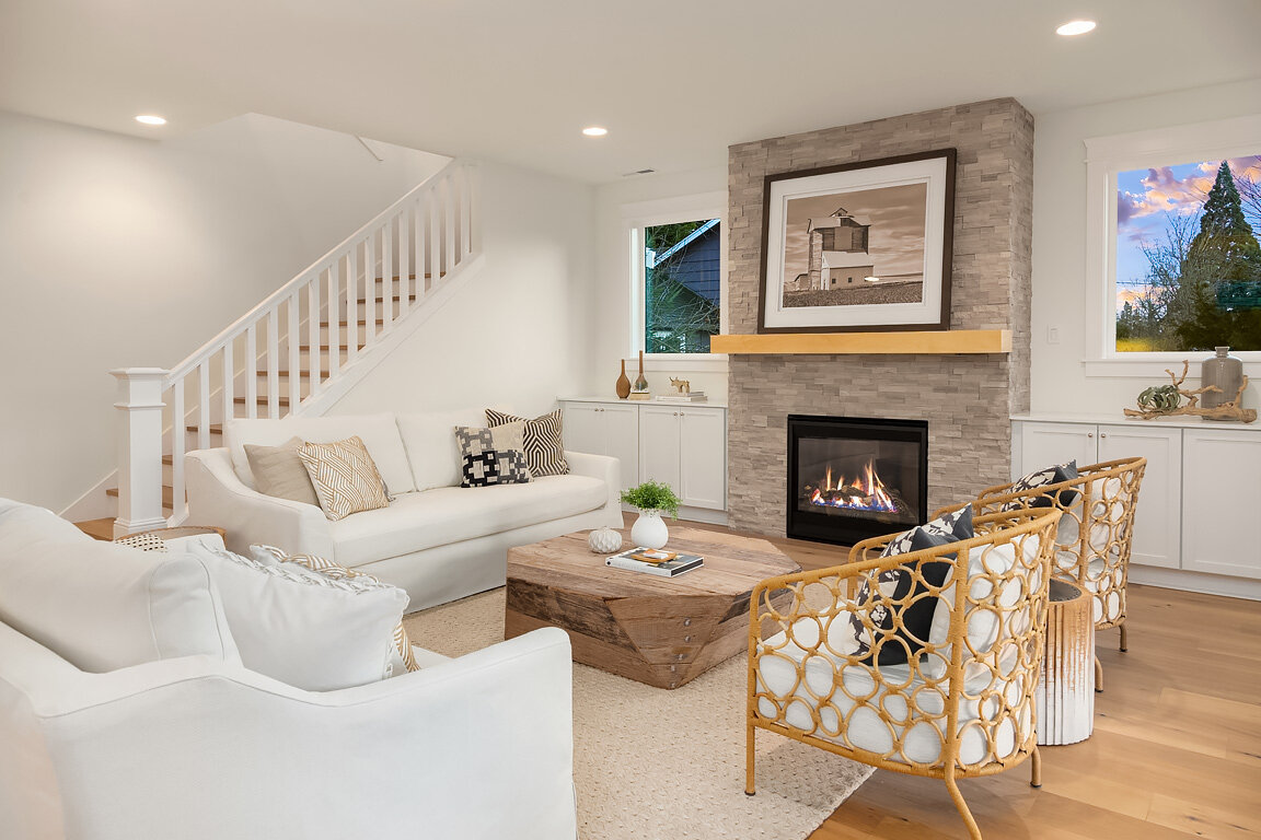 Living room with white sofas, rattan chairs, a wooden coffee table, and a stone fireplace with a framed picture above it. There are side cabinets, vases, and a window showing an outdoor view with trees.