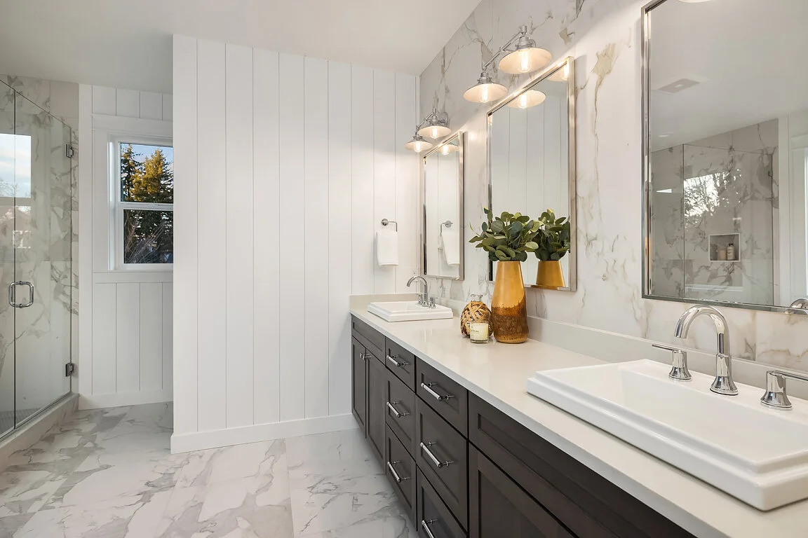 Modern bathroom with double vanity, marble countertops, two mirrors with lights, a vase with greenery, and a walk-in shower with glass door.
