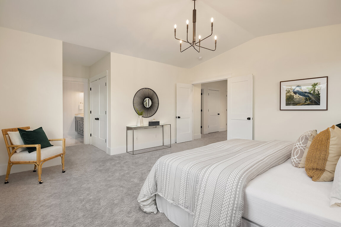 A modern bedroom with a bed, a chair with pillows, a console table, and artwork on the wall, illuminated by a ceiling chandelier.